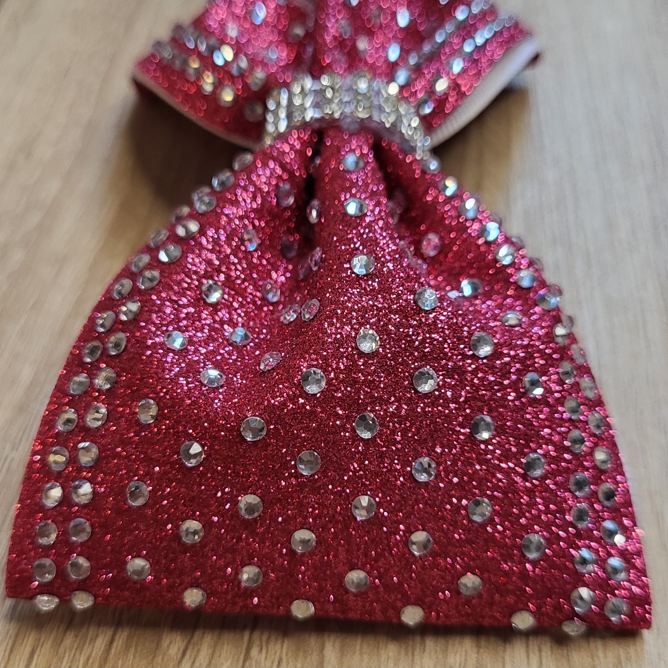 Detailaufnahme einer hochwertigen Cheer Schleife aus pinkem Glitzerstoff. Die Nahaufnahme zeigt die feine Verarbeitung der funkelnden Strasssteine auf dem Bogen sowie das edle, mit Strass besetzte Mittelband, das der Haarschleife Stabilität und extremen Glanz verleiht.