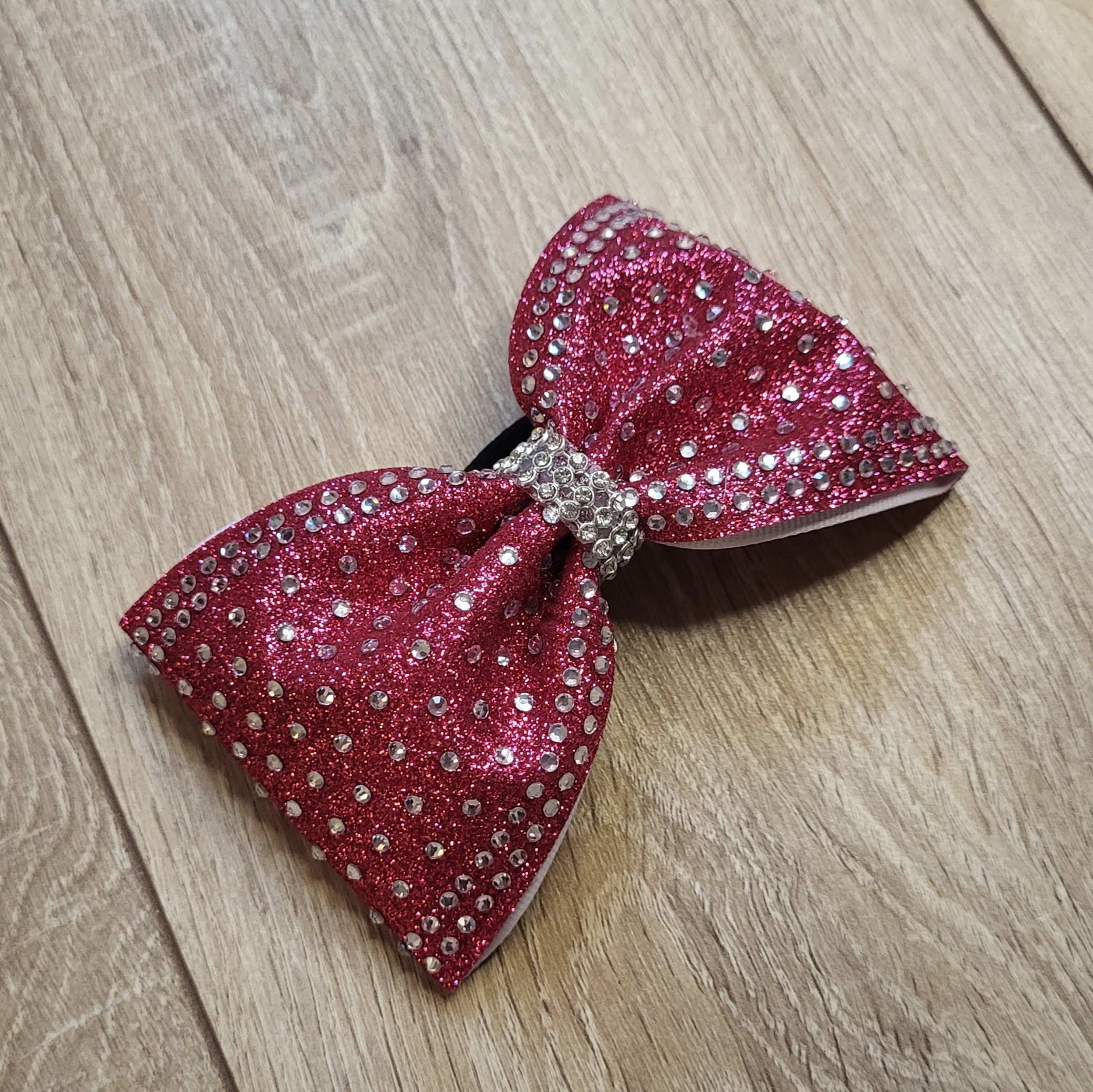 Glitzernder Cheerleading Bow in auffälligem Pink-Rot mit elastischem Haargummi. Die Cheer Schleife zeigt ein exklusives Full-Rhinestone-Design mit präzise gesetzten Strasssteinen und einem funkelnden Center-Band aus Strass (Perspektive leicht schräg).