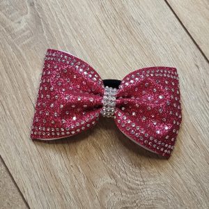 Handgemachte Cheerleader Schleife in funkelndem Zartrot mit feinem Glitzer-Stoff und einem dichten Rand aus funkelnden Strasssteinen. Die Haarschleife ist mit einer edlen Strass-Mitte versehen und liegt flach auf einer hellen Holzoberfläche (Draufsicht). Perfekt für Training und Meisterschaft.